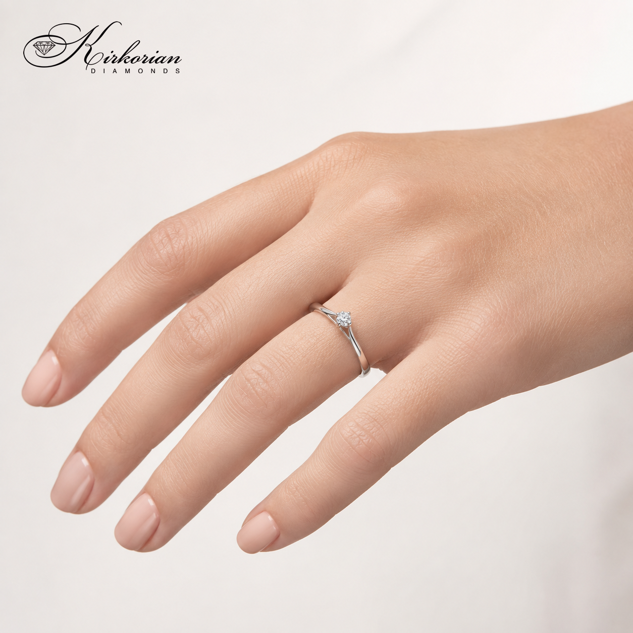 Елегантен годежен пръстен Kirkorian Diamonds K568, носен на безименния пръст на женска ръка с нежен, неутрален маникюр. Деликатният дизайн на бялото злато и блестящият 0.10 ct диамант изглеждат изискано.